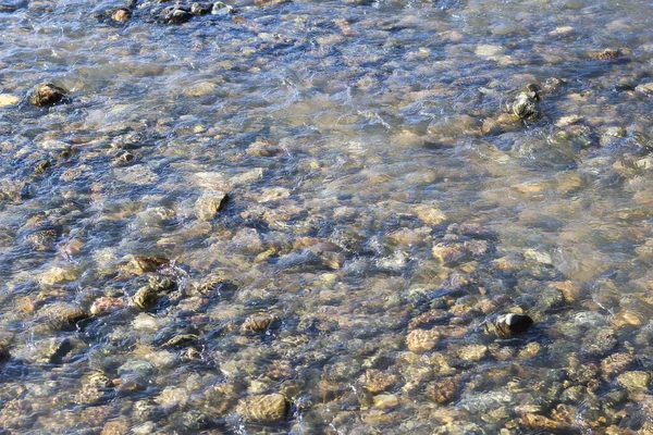 Sakin, sığ nehir akıntısı, kahverengi, yuvarlak çakıl taşlarının üzerinden akıyor. Temiz su dalgalanması, güneş ışığını yansıtan barışçıl bir arka plan oluşturuyor.