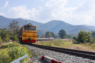 Demiryolu yolculuğundaki nostaljik sarı tren manzaralı kırsal arazide ilerliyor. Barışçıl macera manzarası, engin dağ arkaplanı, güneşli bir günde