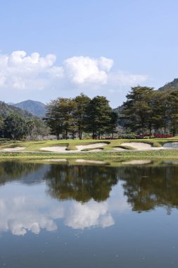 Golf sahasının sakin ve manzaralı manzarasında ağaç ve dağın sudaki yansıması. Temiz mavi gökyüzü ve hiç kimsesi olmayan güzel doğa arka planı
