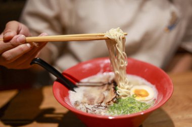 Lezzetli Japon ramen çorbasını domuz ve yumurtalı yiyen kişi. Lokantada doyurucu yemek için kırmızı kasedeki lezzetli erişteyi yemek çubuklarıyla kaldırmak.