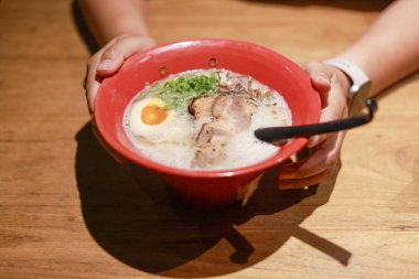 Elinde rahatlatıcı kırmızı kase lezzetli Japon ramen çorbası tutan kişi. Domuz eti ve yumurtalı sıcak yemek tatminkar bir mutfak deneyimi sunuyor.