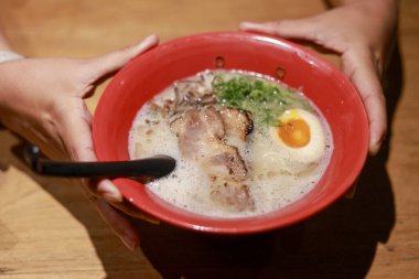 Lezzetli Japon tonkotsu ramen çorbası domuz eti ve kırmızı kasede kaynamış yumurta. Kişi sıcak, tatmin edici tabağı ahşap masanın üzerinde tutar.