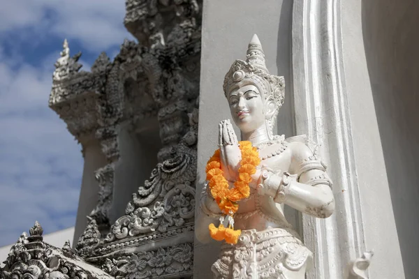 Dini tapınakta dua jesti yapan sakin beyaz Tayland heykeli. Çiçekli bu süslü heykel mavi gökyüzüne karşı barışçıl antik mimariyi gösteriyor.