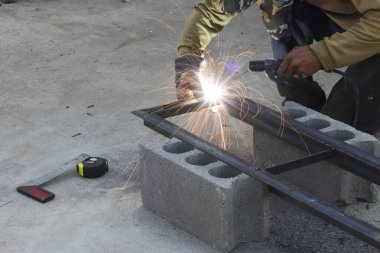 Profesyonel erkek işçi, açık havada inşaat alanında koruyucu eldiven giyerken elinde meşale tutarken parlak kıvılcımlar saçan metal çerçeve yapısına yoğunlaşıyor.