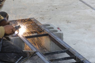 Metal inşaat işçisi parlak kıvılcımlar saçan kaynak aletini tutarken beton zemindeki çelik yapı iskeletini tamir ederken kararlı bir şekilde işe odaklanıyor.
