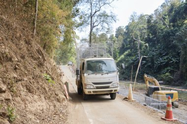 Kırsal yol şantiyesinde ilerleyen beyaz bir kamyon. Ağır makineler ve kazıcı ile dağ yamacının yakınındaki ormanı kazıyor. Güneş ışığı altında.