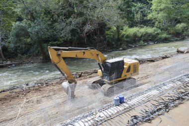 Sarı kazıcı makinası, yeşil ormanın yakınındaki çelik takviye yapısıyla nehir kıyısındaki inşaat sahasında toprak kazıyor.