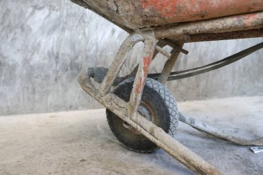 Kirli turuncu el arabası tekerleği ve paslı metal çerçevenin düşük açılı görüntüsünü kapatın. İnşaat alanındaki ham desenli beton arka planda kurumuş beton kalıntılarıyla kaplı.