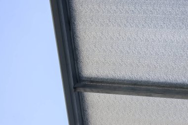 Koyu mavi gökyüzü arka planına karşı koyu kirişlerle çerçevelenmiş dokulu yarı şeffaf polikarbonat çatı panelini gösteren bir yapı.