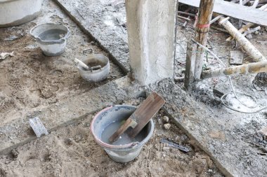 İnşaat alanı alet kovası ve malası ıslak kum zemininde beton sütunla kirli iş dokusunu ve endüstriyel yapı konseptini işçiler olmadan gösteriyor.