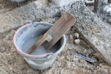 İnşaat alanında su dolu bir kovanın içinde yüzen tahta mala çimento sıva işi için gerekli malzemeyi ve pratik atmosferi gösteriyor.