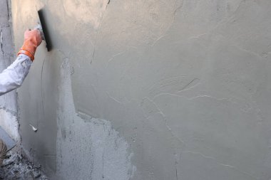 Kapalı inşaat işçisi eli koruyucu turuncu eldiven giyiyor duvar yüzeyine pürüzsüz gri çimento sıva uyguluyor odaklanmış profesyonel efor sarf yapısını onarmak için mala aracı kullanıyor