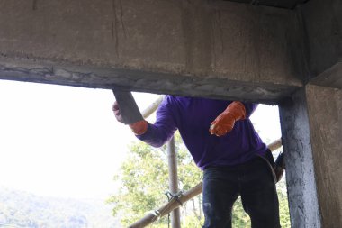 Mor gömlekli inşaat işçisi inşaat alanındaki inşaat iskelesinde mala kullanarak beton kirişi alçılıyor. İşçiliği ve işçiliği gün boyunca özenle gösteriyor.