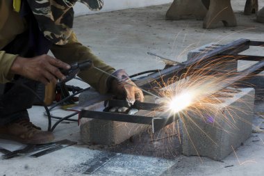 Metal kaynak aleti kullanan profesyonel bir kaynakçı, beton bloktaki çelik inşaat iskeletini odaklanmış duygusal ifadeyle onarırken parlak bir kıvılcım ve flaş yaratıyor.