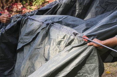 Koyu gri kamp çadırı kuran kişinin yakın görüntüsü açık orman ortamında naylon malzemeyle sakin bir odak ve macera hazırlığı hissediyor.