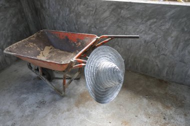 Yapayalnız turuncu el arabası ve geleneksel hasır şapka beton zeminde duruyor. Bitmiş iş hissini ve gri duvar arka planlı inşaat alanında sessiz başarıyı aktarıyor.