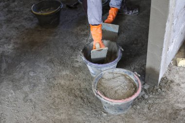 Çalışkan inşaat işçilerinin elleri turuncu eldiven giyip plastik kovanın içine ıslak çimento karıştırarak inşaat sahasında odaklanmış endüstriyel işçiliği gösteriyor.