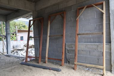 Beton duvarda ahşap kapılı iskeleti olan tamamlanmamış inşaat sahası ham madde ve kum zeminli bina inşaatının ilerlemesini gösteriyor.