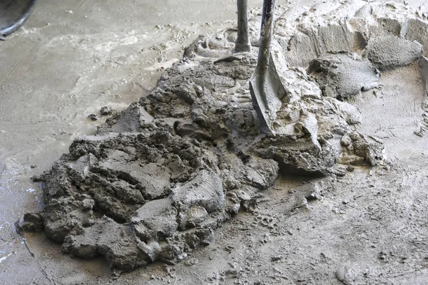 İnşaat manzarasını kapat. Islak gri çimento karışımının zeminde kürekle hazırlandığını, kaba doku ve ağır endüstriyel işlemlerin temel çalışması için hazır olduğunu göster.