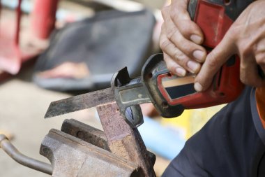 Karşılıklı testereyle çalışan, paslı metal çubuğu mengeneye sıkıştıran odaklanmış bir tamirci. İşçi, atölyede bu amelelik sırasında konsantrasyon gösteriyor