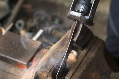 Mengeneye sıkıştırılmış metali kesen elektrikli bir aletle yoğun endüstriyel çalışma. Sürtünmeden çıkan duman ve ısı atölyedeki havayı dolduruyor, ham gücü gösteriyor.