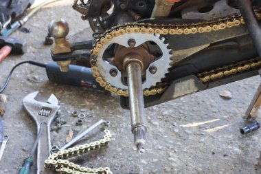 Garajdaki motorsiklet tamiri çalışmalarının yakın görüntüsü. Yerdeki kırık altın zincir, arka dişli, dingil ve metal aletleri gösteren karmaşık mekanik bakım aktivitesi