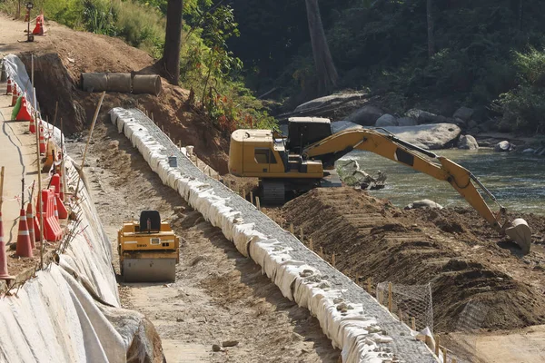 İnşaat alanında kazıcı ve yol silindiri, nehir kıyısında koruma sağlıyor. Bu inşaat mühendisliği projesi kazı, gabion duvarı ve toprak dengeleme çalışmalarını gösteriyor.