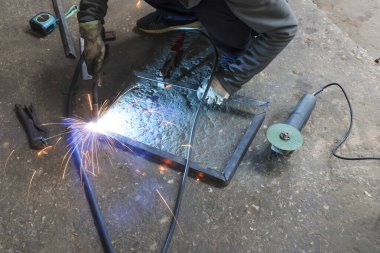 Atölyedeki yetenekli kaynakçı, metal çerçeve kaynak yaparken yoğun odaklanma gösteriyor. Beton zeminde aletlerle endüstriyel süreçten parlak kıvılcımlar uçuşuyor.
