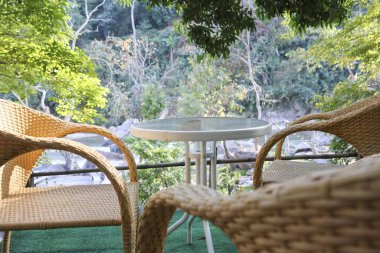 Huzurlu balkonda iki hasır sandalye ve cam masa. Yeşil ormanın güzel doğa manzaralı açık hava mobilyaları. Sessiz zaman için mükemmel bir yer.