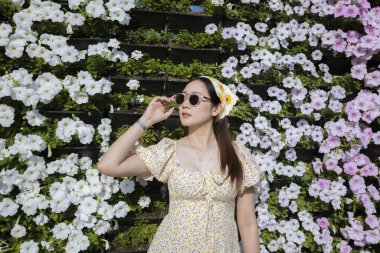 Yazlık elbiseli ve güneş gözlüklü güzel bir Asyalı kadın. Yüzünde sakin bir ifade var. Dikey bahçe duvarı beyaz çiçeklerle dolu şık bir portre.