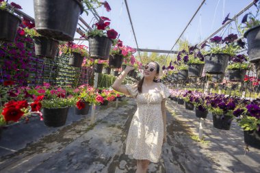 Yazlık elbiseli, neşeli çiçek bahçesinde yürüyüş yapan mutlu güzel kadın. Güneşli bir günde, asılmış petunya saksısıyla dolu renkli serayı keşfediyor.