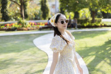 Sarı çiçekli elbisesi ve yeşil parkta yürüyen güneş gözlüklü mutlu, güzel Asyalı kadın. Güneşli bir yaz gününün tadını çıkarıyor. Huzurlu ve mutlu.