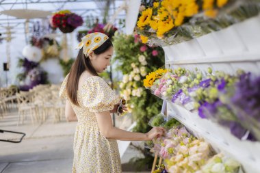 Çiçekçide renkli çiçek buketi seçen, elbise giymiş mutlu genç Asyalı kadın. Bu sakin insan çiçek almak için güzel bir yaşam tarzı alışverişi yapıyor.