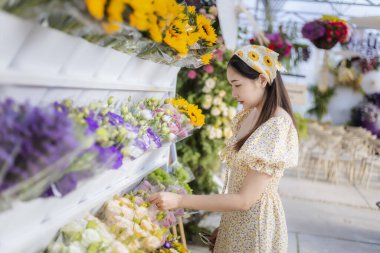 Çiçekçide çiçek buketi seçen sepetli güzel Asyalı bir kadın. Mutlu müşteri taze çiçek pazarında renkli çiçeklere göz atıyor