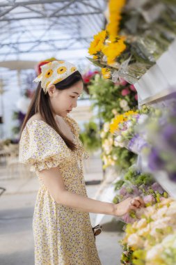 Çiçekçi dükkanından ayçiçeği buketini seçen çiçekçi elbiseli mutlu genç Asyalı kadın. Yaşam tarzı, yerel pazarda taze çiçeklerin tadını çıkarmayı içeriyor.