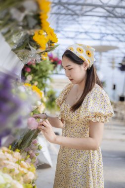 Sarı elbiseli ve saç bantlı sakin Asyalı kadın çiçekçide çiçek buketi seçiyor. Nazik müşteri güzel bir düzenleme için taze çiçek seçer.
