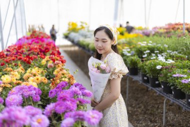 Mutlu genç Asyalı kadın elinde güzel bir buket tutarken büyük bir serada renkli çiçekler alıyor. Huzurlu bir neşe ve hayranlık ifadesi var