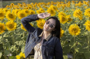 Mutlu gülümseyen kadın ayçiçeği tarlasında güzel bir yaz gününün tadını çıkarıyor. Açık havada canlı sarı çiçeklerin arasında dururken yüzünü parlak güneşten korur.