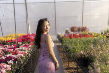Pembe çiçekli elbiseli güzel Asyalı kadın seranın içinde huzurlu bir gülümsemeyle geriye bakıyor. Bu renkli çiçek bahçesi çarpıcı bir arkaplan portresi sunuyor.