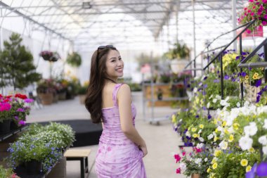 Pembe elbiseli mutlu genç Asyalı kadın çiçek ve çiçek alırken gülümsüyor parlak serada. Neşeli insan bahçe merkezinde eğleniyor.