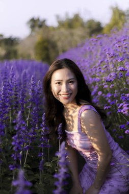 Mutlu gülümseyen Asyalı kadın mor çiçek tarlasında güzel bir günün tadını çıkarıyor. Doğada, dışarıda oturan bu güzel insan, lavanta bahçesinde huzurlu ve neşeli hissediyor.