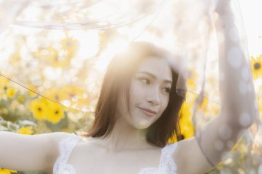 Güzel, genç Asyalı bir kadın, huzurlu bir gülümseme ile güneşli çiçek tarlasında mutlu hissediyor. Saf kumaşı tutuyor, parlak doğada rüya gibi ve barışçıl bir portre oluşturuyor.