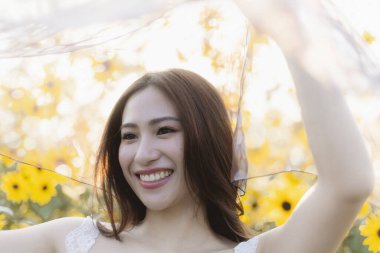 Ayçiçeği tarlasında beyaz peçe tutarken gülümseyen mutlu ve neşeli Asyalı kadın. Sahne, ılık yaz güneşi altında rüya gibi bir his yaratıyor.