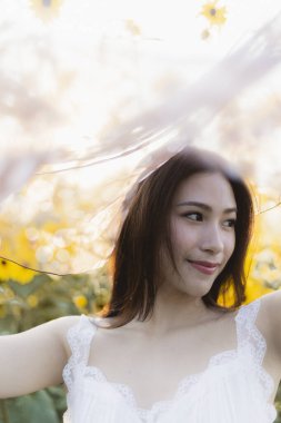 Sarı çiçek tarlasında peçeli beyaz elbiseli mutlu bir Asyalı kadın. Güzel bayan gülümsüyor, doğada güneşli bir yaz gününün tadını çıkarıyor.