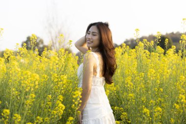 Sarı çiçek tarlasında gülümseyen güzel Asyalı kadın. Mutlu genç yetişkin yaz doğasının tadını çıkarıyor. Sıcak güneş ışığı altında huzurlu ve tasasız hissediyor.