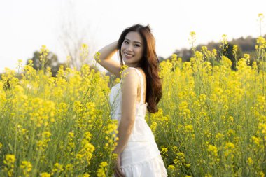 Yaz boyunca sarı çiçek tarlasında mutlu Asyalı kadın portresi. Beyaz elbiseli neşeli genç bayan doğanın tadını çıkarıyor, kaygısız bir ifadeyle gülümsüyor.