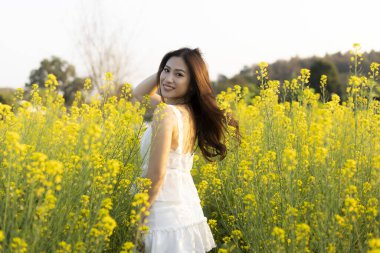 Beyaz elbiseli mutlu Asyalı kadın sarı çiçek tarlasında dönüyor. Güzel bir genç portresi kaygısız yaz gününün tadını çıkarıyor doğayla iç içe.