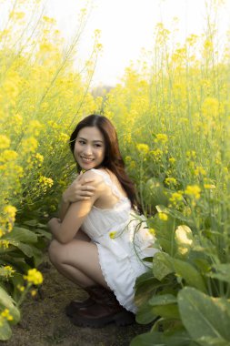 Sarı çiçek tarlasında gülümseyen mutlu Asyalı kadın. Bu güzel kız güneşli bahar gününde doğanın tadını çıkarıyor. Canlı kolza tohumu çiçeklerinin arasında çömeliyor.