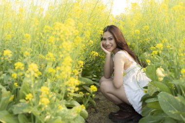 Güzel sarı çiçek tarlasında gülümseyen mutlu genç Asyalı kadın. Beyaz elbiseli hoş bayan, sakin bir yaz gününün tadını çıkarıyor.