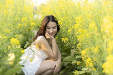 Sarı çiçek tarlasında çömelmiş mutlu gülümseyen güzel Asyalı kadın. Neşeli kadın, sakin yaz gününde, parlak güneş ışığında doğanın tadını çıkarıyor.